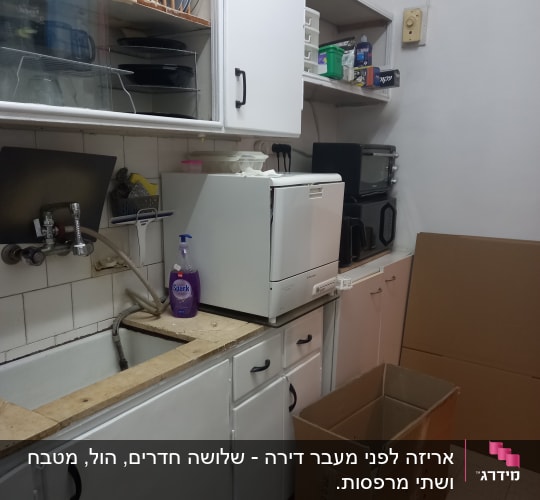 מטבח עם ארונות לבנים, קרטון פתוח, כיור וחומרי ניקוי.